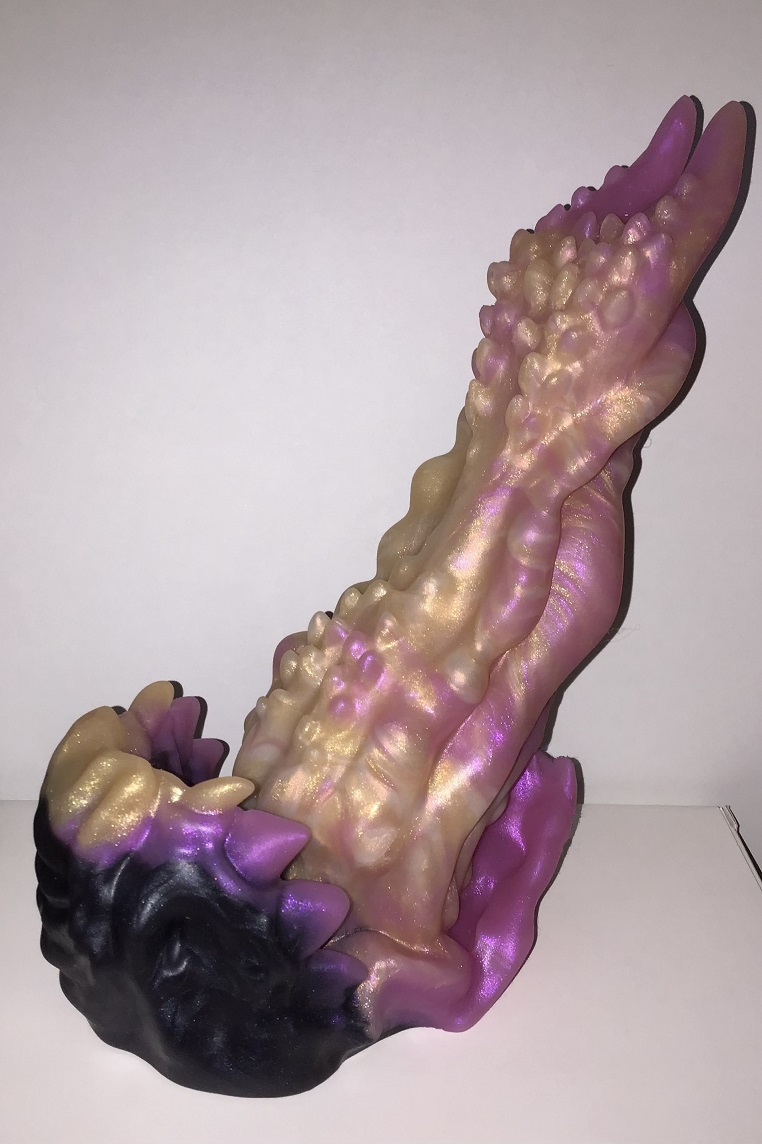 The base is an alien maw with teeth, opened wide. The shaft is a muscular tongue with spikes along the top side. The tongue is a shimmery marble of pink, gold, and pale gold. The base includes the same colors, but the top half of the jaw is mostly black.